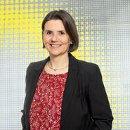 Porträtfoto von einer Frau mit kinnlangen braunen Haaren, die ein rotes Shirt mit weißem Muster und einen schwarzen Balzer trägt und vor einer gelben-weißen Glaswand steht.