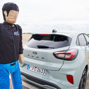 Das Bild zeigt einen Crashtest-Dummy hinter einem grauen Auto.