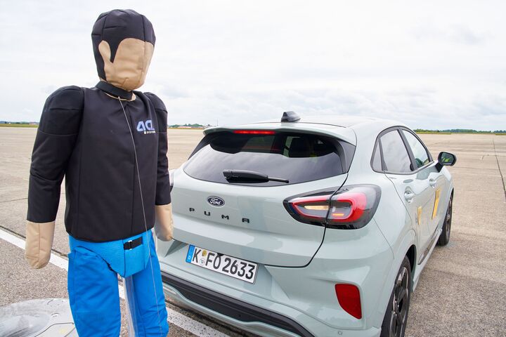 Das Bild zeigt einen Crashtest-Dummy hinter einem grauen Auto.