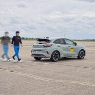 Das Bild zeigt einen zu Fuß gehenden Crashtest-Dummy, der sich hinter einem grauen Auto vorbeibewegt. Die Bewegung wird mittels einer Serienaufnahme dargestellt. 