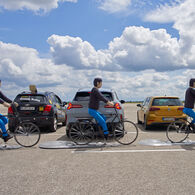 Notbremsassistenten für die Rückwärtsfahrt im ÖAMTC-Test Das Bild zeigt drei Crashtest-Dummies auf Fahrrädern. Sie fahren hintereinander hinter drei Autos vorbei, die nebeneinander und im rechten Winkel zur Fahrtrichtung der radfahrenden Dummies parken. Das mittlere, graue Auto parkt gerade aus, der mittlere der drei radfahrenden Dummies befindet sich  genau hinter dem Fahrzeug. Die Bremslichter leuchten rot.