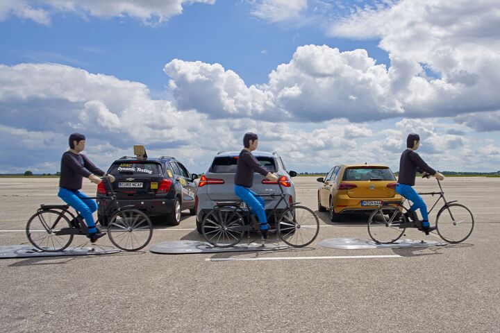 Das Bild zeigt drei Crashtest-Dummies auf Fahrrädern. Sie fahren hintereinander hinter drei Autos vorbei, die nebeneinander und im rechten Winkel zur Fahrtrichtung der radfahrenden Dummies parken. Das mittlere, graue Auto parkt gerade aus, der mittlere der drei radfahrenden Dummies befindet sich  genau hinter dem Fahrzeug. Die Bremslichter leuchten rot.