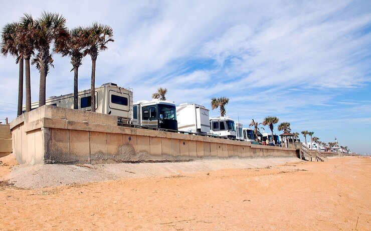 Camping am Strand Mehrere Wohnmobile, die nebeneinander gereiht an einer palmengesäumten Strandpromenade stehen. Im Vordergrund ist ein feiner Sandstrand zu sehen.