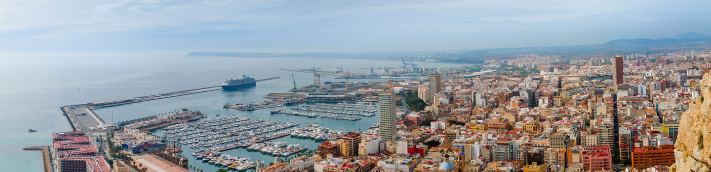 Alicante präsentiert sich als charmante Küstenstadt, deren Silhouette sich mit pastellfarbenen Gebäuden und einem leuchtenden Hafen gegen den weiten, blauen Himmel abzeichnet.