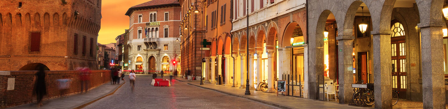 Ein ruhiger Blick auf eine Straße in Ferrara in der Abenddämmerung, mit den sanften Lichtstrahlen, die die historischen Gebäude und gepflasterten Straßen in warmes Licht tauchen.