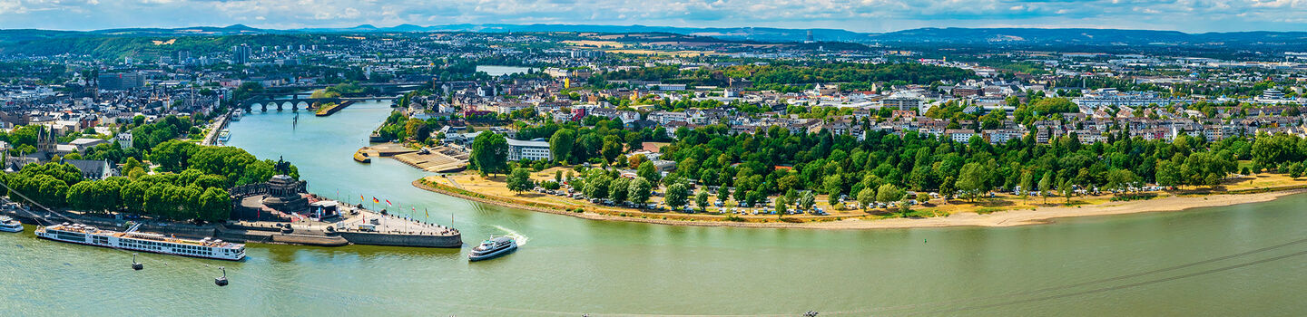 Zusammenfluss von Rhein und Mosel in Koblenz, mit den Städten im Hintergrund, während die Flüsse unter einem strahlend blauen Himmel aufeinandertreffen.