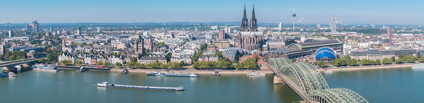 Panorama von Köln, mit dem Rhein, der Brücke und dem Kölner Dom im Hintergrund, alles unter einem klaren blauen Himmel.