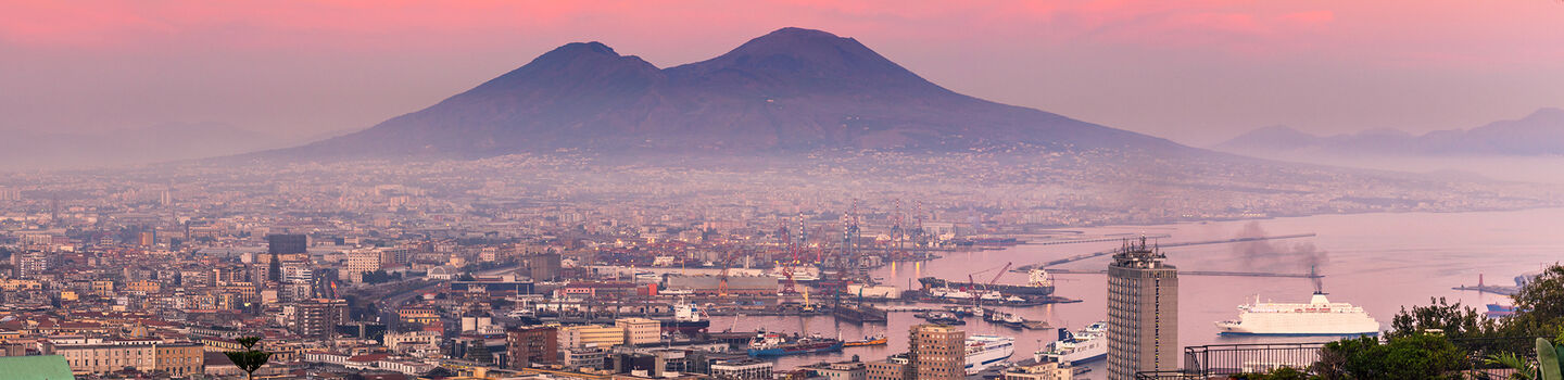 Ein malerischer Blick zeigt Neapel und den imposanten Vesuv im rosa Licht des Sonnenuntergangs, während die Stadt in sanften, warmen Farben erstrahlt und der Vulkan majestätisch über der Szenerie thront.