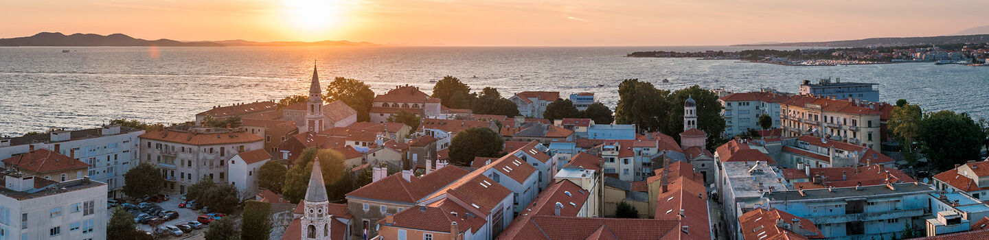 Blick auf Zadar bei Sonnenuntergang, mit den roten Dächern der Stadt und dem Meer, das in warmes Licht getaucht ist.