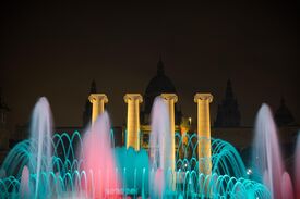 Farbenprächtige nächtliche Wasserspiel-Show mit türkisfarbenen Wasserbögen und rosavioletten Fontänen vor golden leuchtenden Säulen und einem schwach beleuchteten historischen Gebäude, was eine dramatische, festliche Stimmung erzeugt.