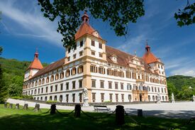 Außenaufnahme des Schloss Eggenberg bei Graz. In das Bild ragen die grünen Äste eines Baumes im Schlosspark, hinter dem Schloss grüne Wälder bei blauem Himmel.