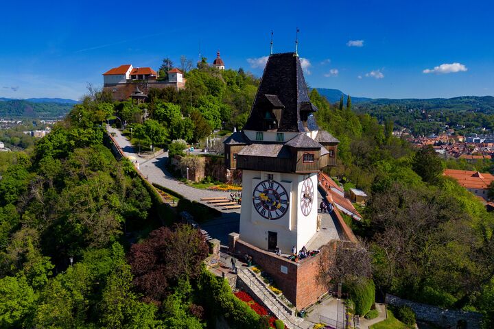 Das Wahrzeichen von Graz, der Grazer Uhrturm, im Vordergrund. Im Hintergrund erstreckt sich der grün bewachsene Schlossberg bei blauem Himmel.