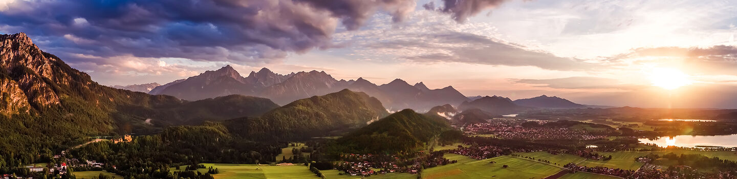 Malerisches Panorama des Forggensees und Schwangaus im Allgäu, mit dem weltberühmten Schloss Neuschwanstein im Hintergrund, umrahmt von den sanften Hügeln des Allgäus und einem warmen, goldenen Sonnenaufgang.