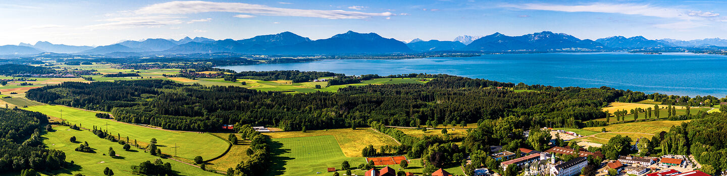 Luftpanorama des Chiemsees, umgeben von grünen Hügeln und den Alpen im Hintergrund, mit dem glitzernden Wasser des Sees, das im Sonnenlicht schimmert.