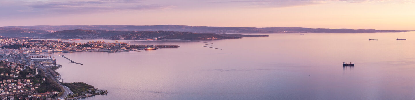 Aus großer Entfernung zeigt sich die schöne Stadt Triest im warmen Licht des Sonnenuntergangs, umgeben von sanften Hügeln und dem weiten Meer.