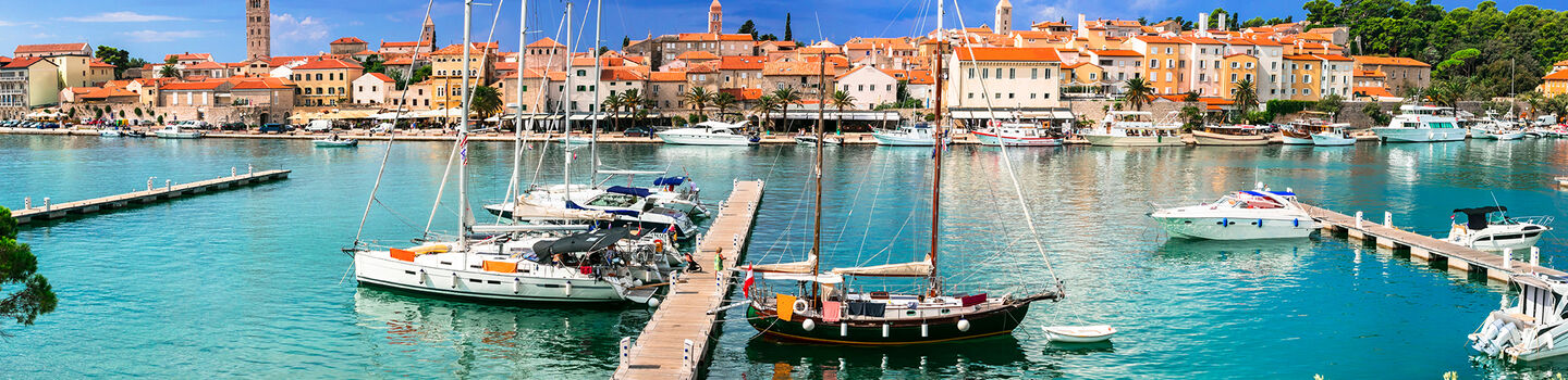 Blick auf die Altstadt von Rab, mit dem Hafen im Vordergrund, dem azurblauen Wasser und den historischen Gebäuden der Stadt im Hintergrund.