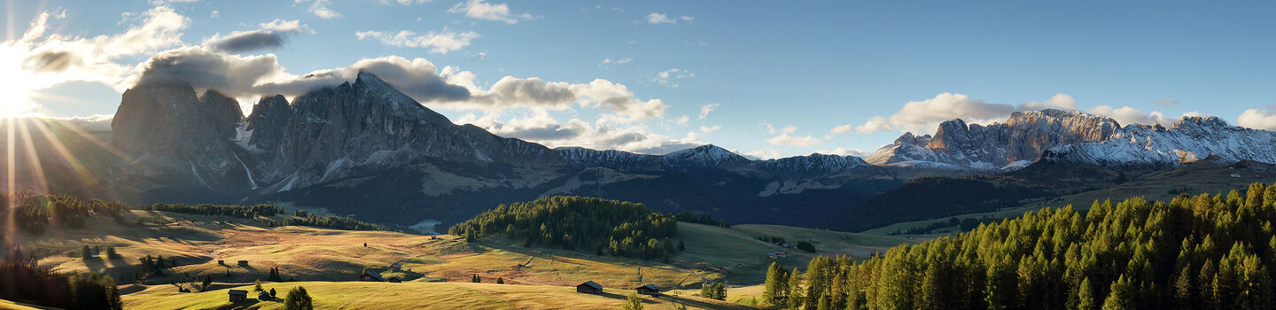 Ein atemberaubender Blick über die Seiser Alm in Südtirol, mit den beeindruckenden Dolomiten im Hintergrund und der Sonne, die hinter den Bergen hervorstrahlt.