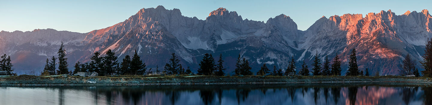 Die Sonne senkt sich über dem Wilden Kaiser Massiv und hüllt das ehrfürchtige Bergpanorama in klar schimmernde rosa-lila Töne der Dämmerung.