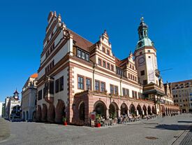 Das historische alte Rathaus mit aufwendiger Architektur, darunter Bögen und ein hoher Uhrturm. Straßencafé mit Gästen unter strahlend blauem Himmel.