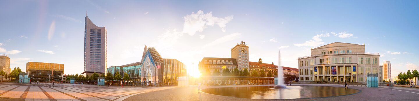 Panoramablick auf den Leipziger Augustusplatz bei Sonnenuntergang mit modernen und historischen Gebäuden, einem Brunnen und einem Kopfsteinpflasterplatz. Helle, ruhige Atmosphäre.
