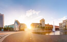 Panoramablick auf den Leipziger Augustusplatz bei Sonnenuntergang mit modernen und historischen Gebäuden, einem Brunnen und einem Kopfsteinpflasterplatz. Helle, ruhige Atmosphäre.