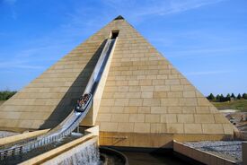 Ein Boot gleitet von einer großen Pyramidenkonstruktion im Belantis Freizeitpark eine Wasserrutsche hinunter, umgeben von einem Spritzer Wasser, was unter einem klaren blauen Himmel Begeisterung auslöst.