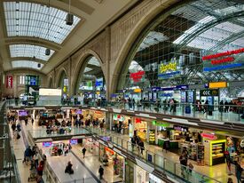 Innenansicht des Leipziger Hauptbahnhofs mit gewölbten Decken und großen Glasfenstern. Zahlreiche Menschen flanieren an Geschäften und Restaurants vorbei und schaffen eine lebhafte Atmosphäre.