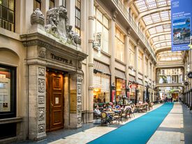 Die prachtvolle Mädler Passage mit einem kunstvoll verzierten Steineingang, der mit „TREPPE A“ beschriftet ist. Helles Tageslicht, lebhafte Geschäfte und Menschen, die an Tischen im Freien speisen, vermitteln eine lebhafte Atmosphäre.