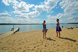 Zwei Personen in Sommerkleidern stehen an einem Sandstrand unter blauem Himmel mit flauschigen Wolken. Ein Steg ragt in das ruhige Wasser des Markkleeberger Sees bei Leipzig hinein.