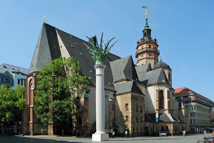 Die historische Nikolaikirche in Leipzig mit hohem Glockenturm erhebt sich vor strahlend blauem Himmel. Im Vordergrund ist eine Säule mit palmenartiger Spitze zu sehen. Menschen gehen in der Nähe entlang.