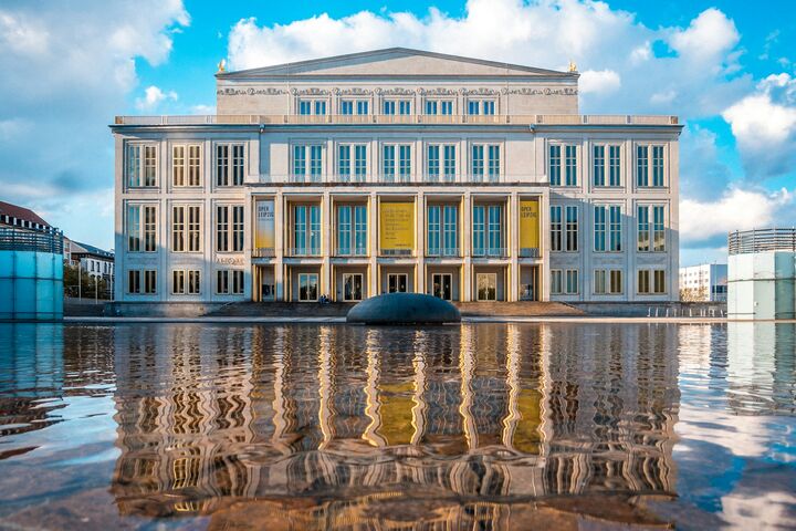 Die elegante neoklassizistische Oper von Leipzig mit großen Fenstern und Säulen spiegelt sich in der ruhigen Wasseroberfläche. Im Hintergrund strahlend blauer Himmel und weiße Wolken.