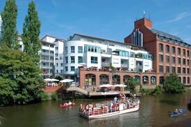 Die Uferpromenade im Leipziger Stadtteil Plagwitz ist geprägt von modernen Gebäuden aus weißen und roten Backsteinen, darunter ein Restaurant direkt am Wasser. Die Menschen genießen Bootsfahrten; die Atmosphäre ist lebhaft und sommerlich.