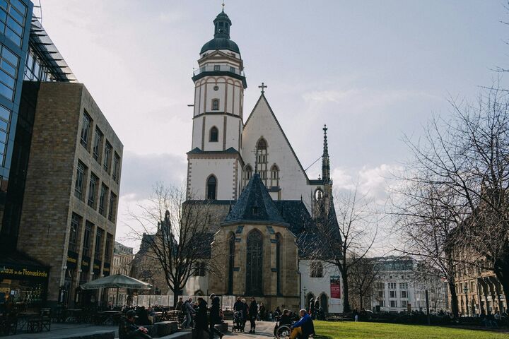 Die Thomaskirche in Leipzig mit hohem Turm, umgeben von kahlen Bäumen und modernen Gebäuden. Auf dem sonnigen Platz haben sich Menschen versammelt.