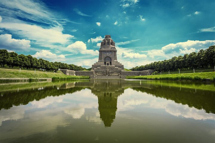Das Völkerschlachtdenkmal in Leipzig, ein imposantes Steinmonument, spiegelt sich in einem ruhigen Teich, umgeben von üppigem Grün und unter einem strahlend blauen Himmel mit vereinzelten Wolken – eine Oase der Ruhe.