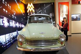 In einem schwach beleuchteten Raum im Zeitgeschichtlichen Forum Leipzig steht ein hellgrüner Trabi im Mittelpunkt. Ein Mann und eine Frau lesen daneben Informationsmaterial; im Hintergrund sind Wandmalereien mit urbanen Motiven zu sehen.