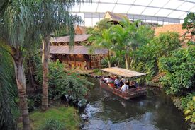 Ein kleines Boot mit Passagieren gleitet über einen üppigen, tropischen Indoor-Fluss in der Gondwanaland Tropenerlebniswelt. Palmen und sattes Grün umgeben ein rustikales Holzgebäude in der Nähe.