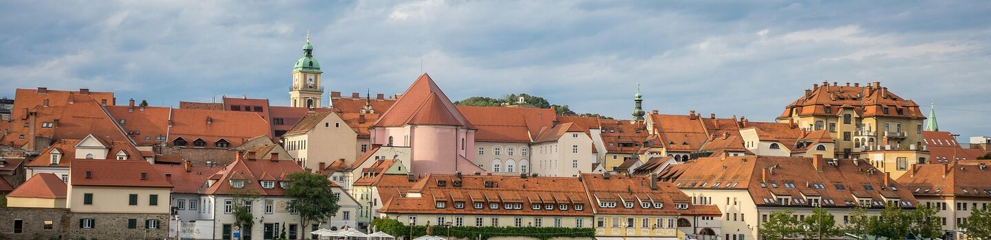 Das malerische Flussufer der Drau in Maribor mit rotgedeckten Gebäuden unter bewölktem Himmel, die sich im Wasser spiegelt, vermittelt eine ruhige, historische europäische Atmosphäre.