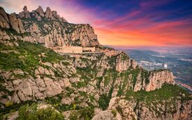 Die schroffen Klippen von Montserrat und ein Kloster überblicken ein fernes Tal unter einem leuchtend rosa-orangefarbenen Sonnenuntergang – ein Anblick, der zugleich friedvoll und dramatisch wirkt.