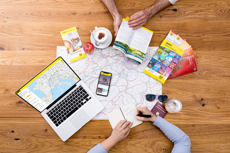Zwei Personen planen eine Reise, nutzen Karten, Reiseführer, einen Laptop und ein Smartphone.
