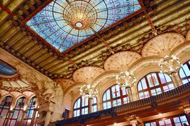 Prunkvolle historische Halle mit einem leuchtenden Buntglas-Oberlicht, geschnitzten Steinsäulen, Kronleuchtern und hohen Bogenfenstern; das imposante Interieur wirkt warm und opulent.