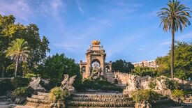 Ein kunstvoller Steinbrunnen und ein imposanter Torbogen in einem üppigen Park in Barcelona, ​​eingerahmt von Palmen und Grün unter einem strahlend blauen Himmel; die Szene wirkt friedvoll und majestätisch.