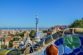 Eine farbenfrohe Mosaikbank bietet einen Blick über die Dächer Barcelonas und das blaue Mittelmeer, während sich in der Ferne unter klarem Himmel ein Turm im Stil Gaudís erhebt. Die Szene wirkt hell und friedlich.