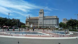 Ein sonniger Stadtplatz mit einem imposanten historischen Gebäude, einem Brunnen, flanierenden Besuchern und Tauben unter blauem Himmel; die Szene wirkt lebendig und offen.