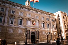 Sonnenlicht beleuchtet ein imposantes, historisches Regierungsgebäude aus Stein mit Rundbogenfenstern, Balkonen sowie spanischen und regionalen Flaggen, während sich darunter Fußgänger bewegen – ein Anblick, der bürgerlichen Stolz vermittelt.