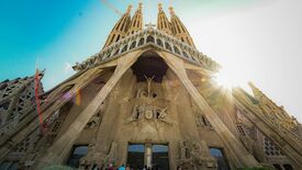 Ansicht der Sagrada Família in Barcelona aus der Froschperspektive: Detailreiche Steinmetzarbeiten und hoch aufragende Türme, die im Gegenlicht der Sonne vor blauem Himmel erstrahlen und eine ehrfurchtgebietende Atmosphäre schaffen.