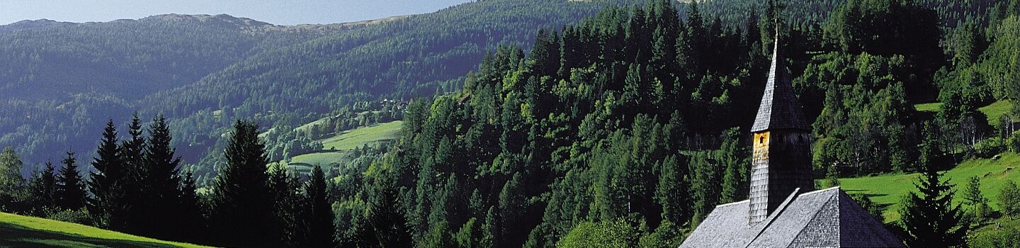 Kleine Kapelle auf einer grünen Wiese, dahinter erhebt sich ein bewaldeter Berg