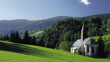 Kleine Kapelle auf einer grünen Wiese, dahinter erhebt sich ein bewaldeter Berg