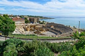 Die antiken Ruinen des Amphitheaters in Tarragona mit geschwungenen Steinsitzreihen überblicken ein ruhiges, blaues Meer; Gärten, Gebäude der Ortschaft und ein strahlender Himmel bilden eine friedliche Szenerie.