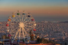 Ein hell erleuchtetes Riesenrad überblickt bei Sonnenuntergang eine weitläufige Küstenstadt; seine bunten Gondeln leuchten warm vor dem Hintergrund des fernen Meeres und des diesigen Himmels.