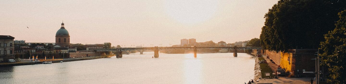 Sonnenuntergang an der Garonne in Toulouse, an deren Uferpromenade gemächlich Menschen spazieren und sitzen. Die Silhouette einer Kuppel und einer Brücke in der Ferne unterstreicht die friedliche Szenerie.